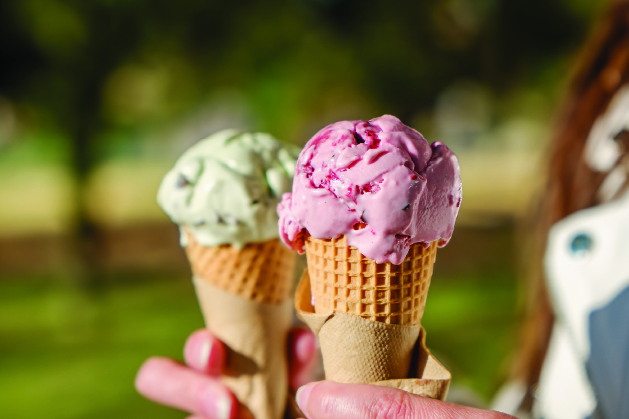 Two waffle cones held in hands outdoors: foreground cone with a scoop of pink berry ice cream with visible fruit pieces beginning to melt, background cone with pale green mint chip ice cream, both wrapped in paper sleeves against a blurred sunny park backdrop.